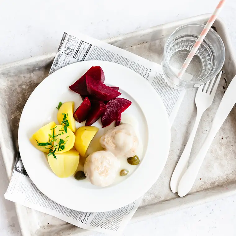 Ein Teller mit Königsberger Klopsen, Kartoffeln und Rote-Bete-Scheiben, daneben ein Glas Wasser.