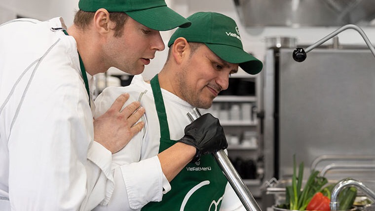 Zwei Köche in grünen Schürzen arbeiten konzentriert an einem großen Kochtopf in der Küche.