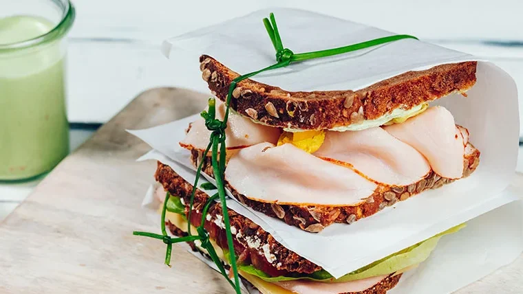 Zwei Vollkorn-Sandwiches mit Putenbrust, Salat und Käse, verpackt in Pergamentpapier und gebunden mit Schnittlauch.